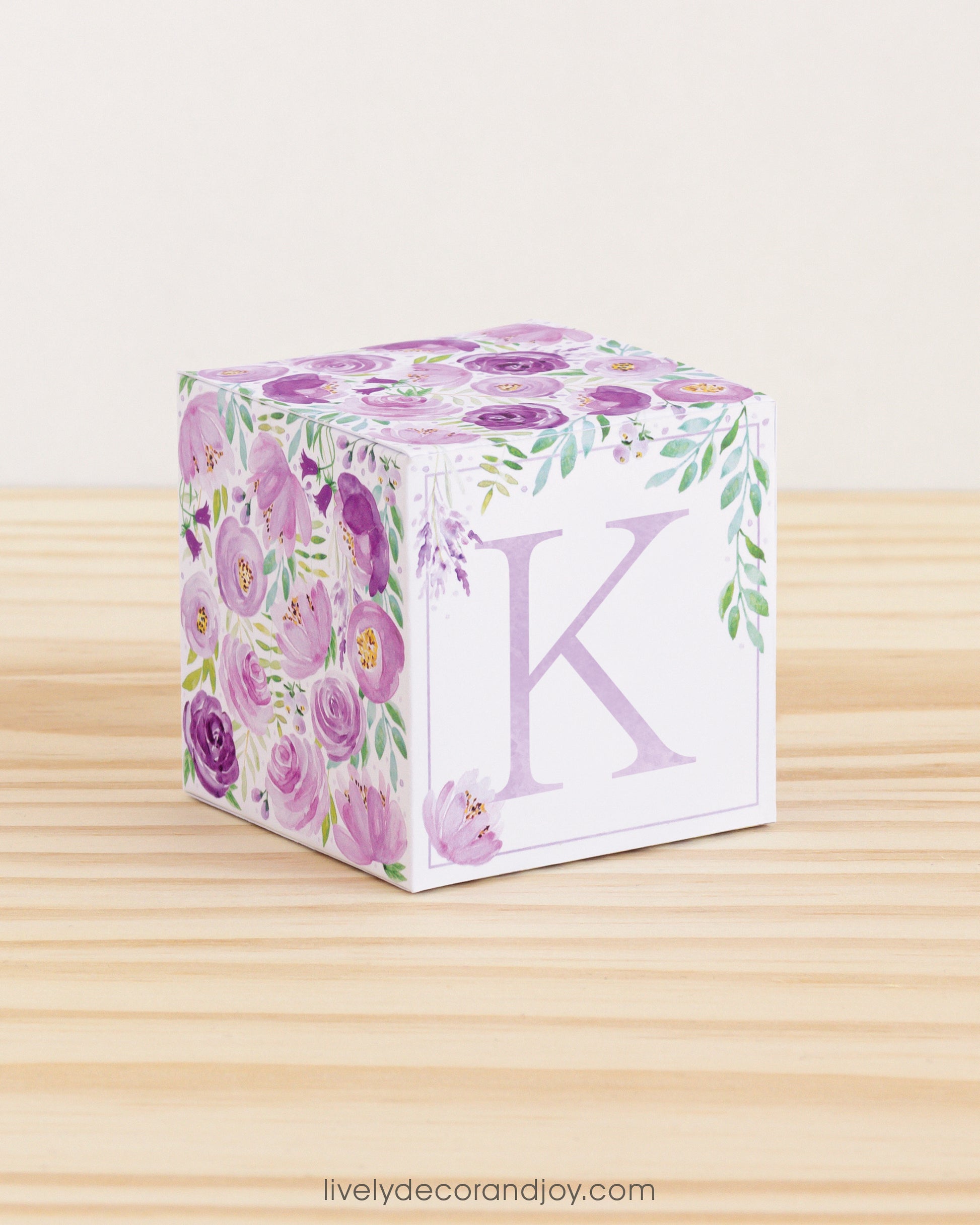 Big DIY block with the letter K in lavender and floral designs in purple and green.The paper cube is on a wooden table.