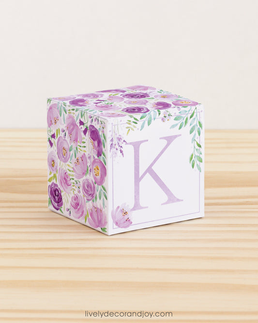 Big DIY block with the letter K in lavender and floral designs in purple and green.The paper cube is on a wooden table.