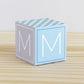 5 minute craft, ABC block on a wooden surface. Made of paper. On the sides, it has the capital M.