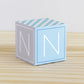 On a wooden table is a paper building block in tints of blue and light grey. Has the letter N on the sides.