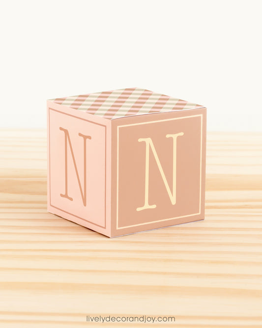 On a wooden table is a paper building block in farmhouse colors. Has the letter N on the sides.