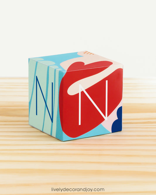 On a wooden table is a paper building block with abstract designs. Has the letter N on the sides.
