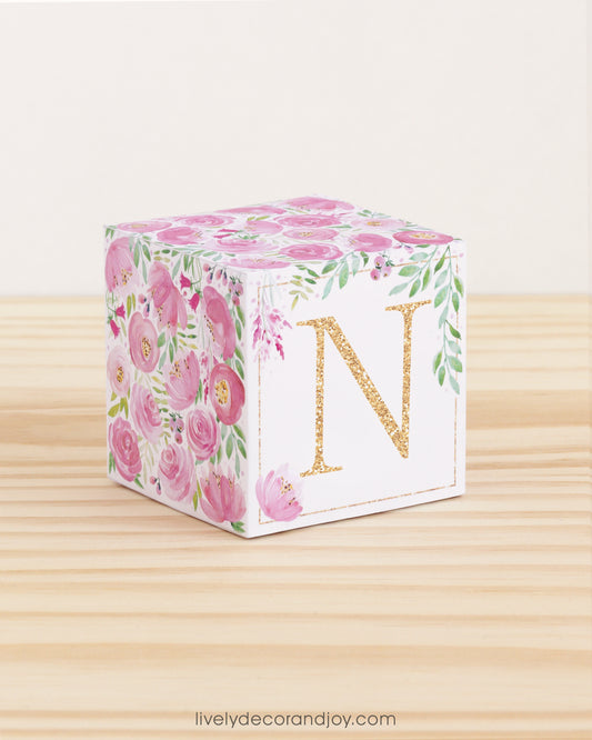 On a wooden table is a paper building block with pink and green floral designs and the letter N in gold on the front.