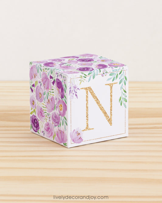On a wooden table is a paper building block with purple and green floral designs and the letter N in gold on the front.