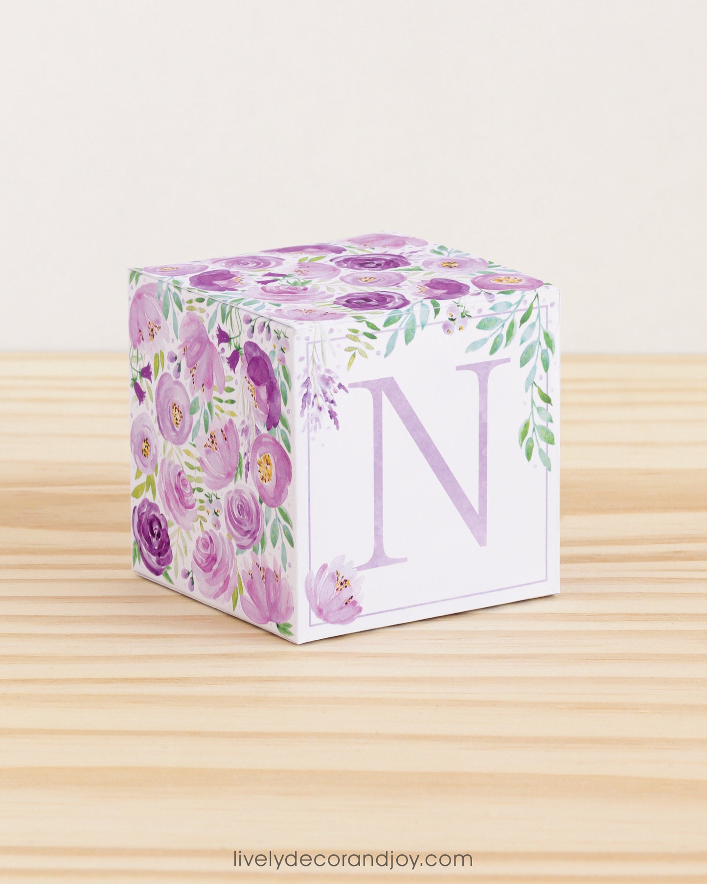 On a wooden table is a paper building block with purple and green floral designs and the letter N in lavender on the front.