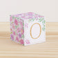 Baby block with blush and lavender floral designs and a peach letter O is on a wooden surface. It is a paper building block.