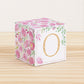 A baby block with pink and green floral designs and a gold letter O is on a wooden surface. It is a paper building block.