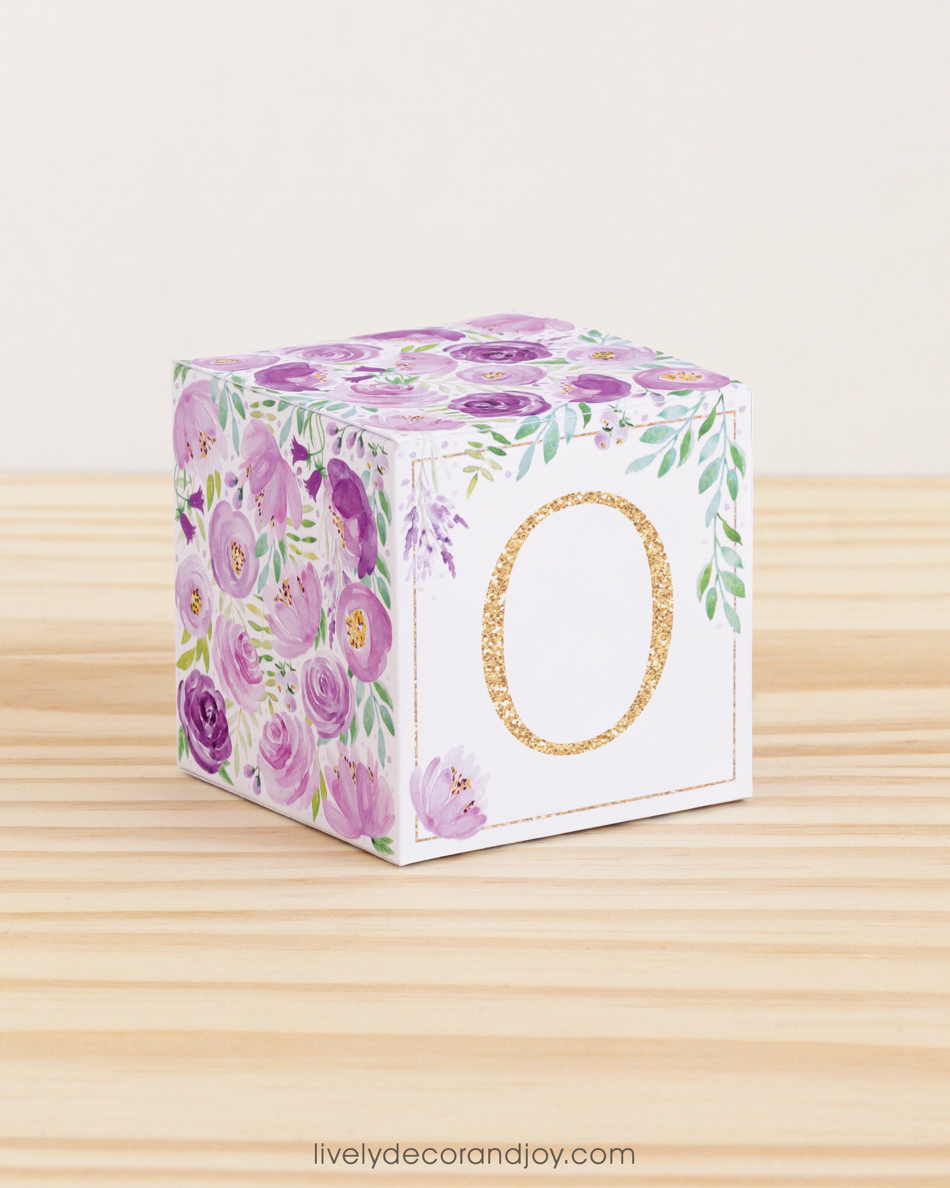 A baby block with purple and green floral designs and a gold letter O is on a wooden surface. It is a paper building block.