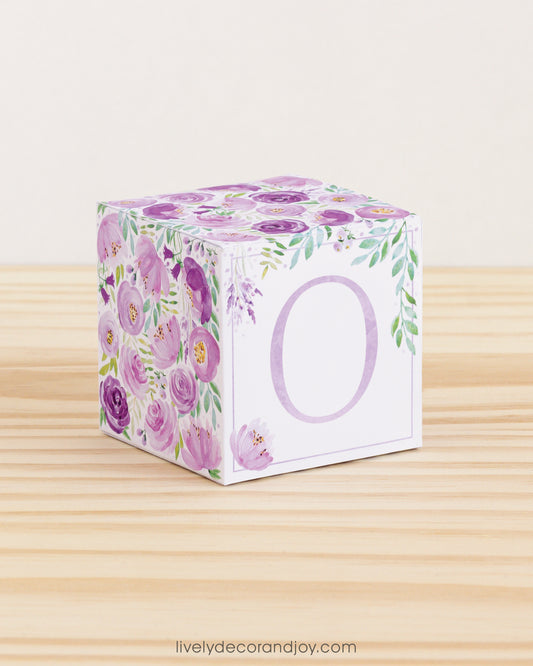Baby block with purple and green floral designs and a lavender letter O is on a wooden surface. It is a paper building block.