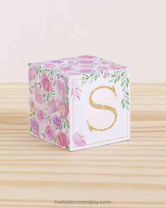 Building block made of printed paper with floral cottagecore designs and the letter S on the front. It is on a wooden table.