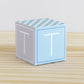 Paper building block in tints of blue and light grey. It has the letter T on the sides and is on a wood table or shelf.