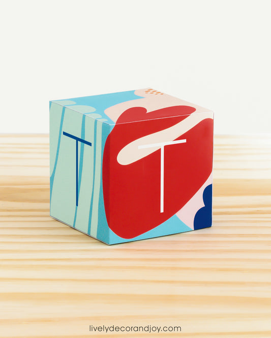 Paper building block in coral reef colors. It has the letter T on the sides and is on a wood table or shelf.