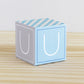 A baby block in tints of blue and light grey with the letter U. It is on a wooden surface and is made of printed paper.