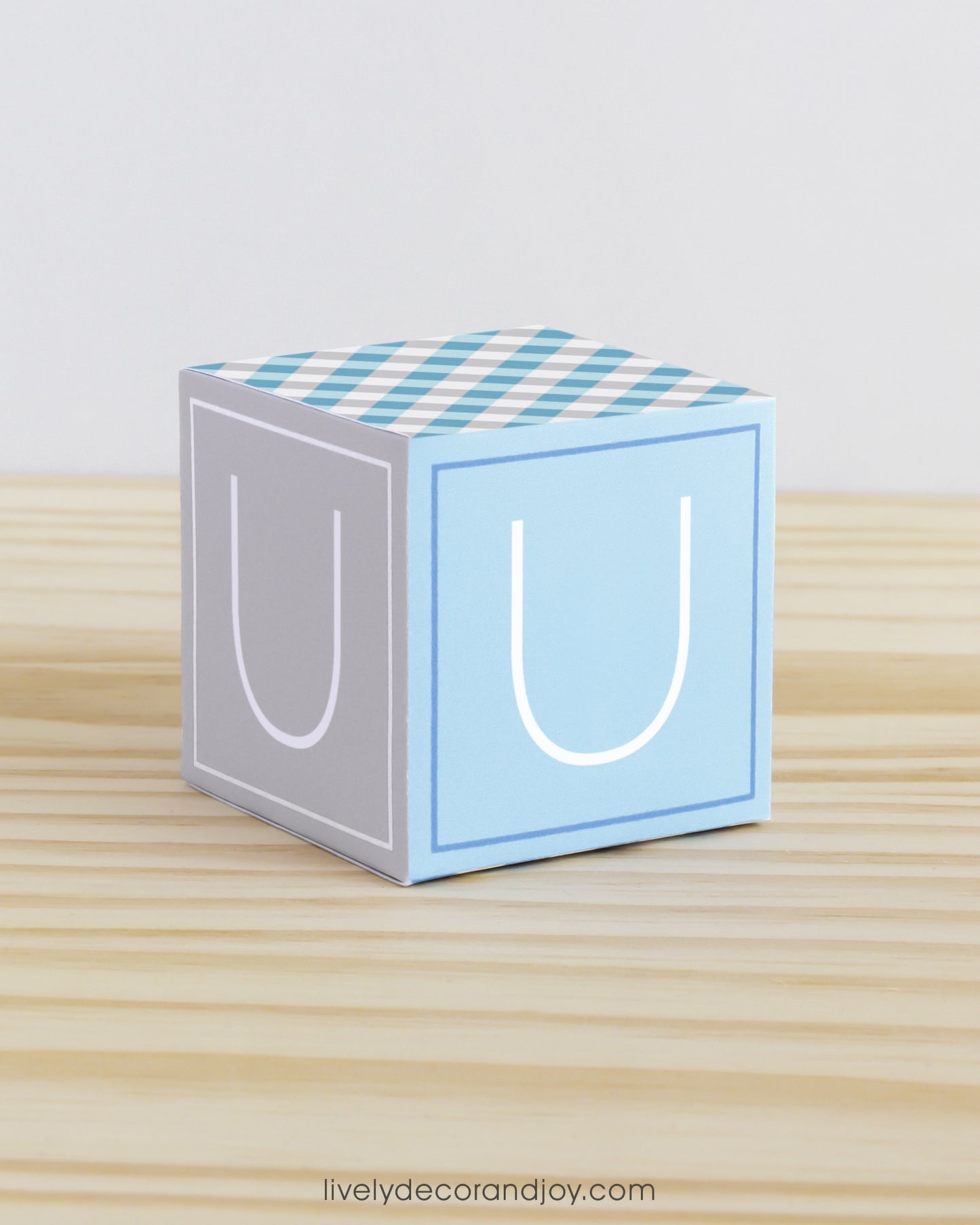 A baby block in tints of blue and light grey with the letter U. It is on a wooden surface and is made of printed paper.