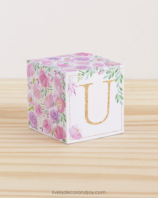 A baby block with blush and lavender floral designs and a peach letter U on a wooden surface. It is a paper building block.
