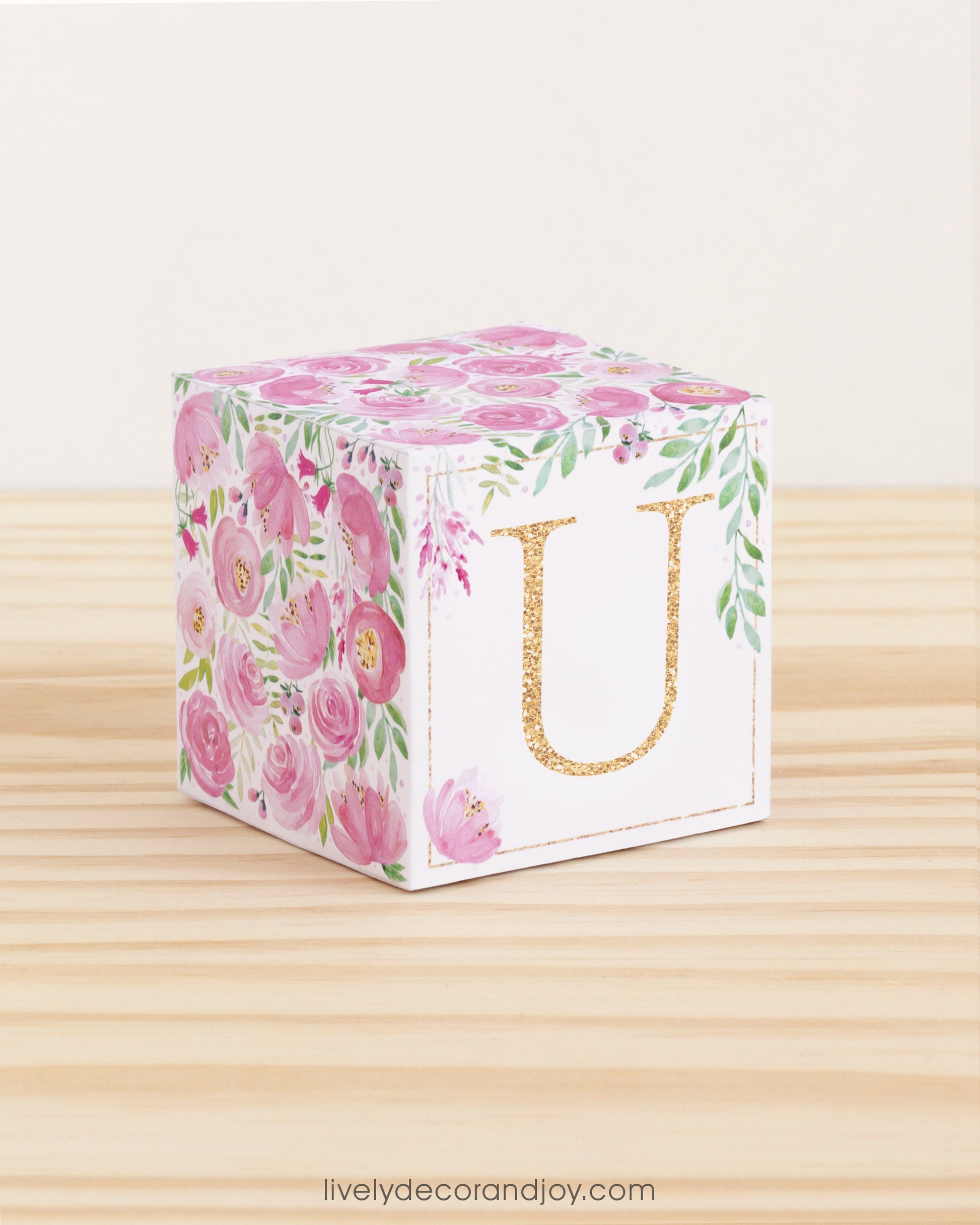 A baby block with pink and green floral designs and a gold letter U is on a wooden surface. It is a paper building block.