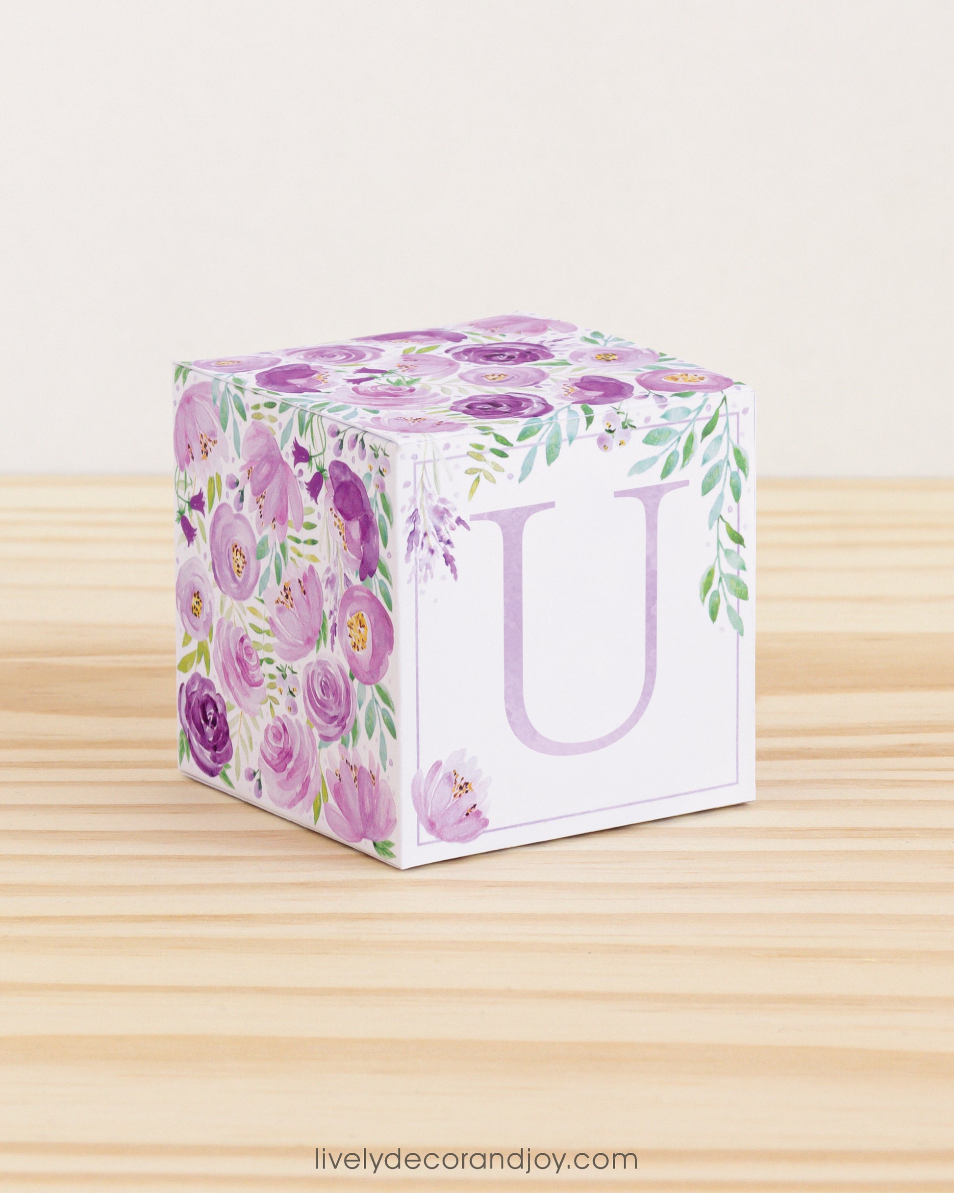 A baby block with purple and green floral designs and a lavender letter U on a wooden surface. It is a paper building block.
