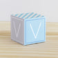 Paper building block in tints of blue and light grey. It has the letter V on the sides and is on a wood table or shelf.