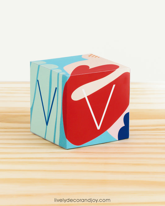 Paper building block in coral reef colors. It has the letter V on the sides and is on a wood table or shelf.
