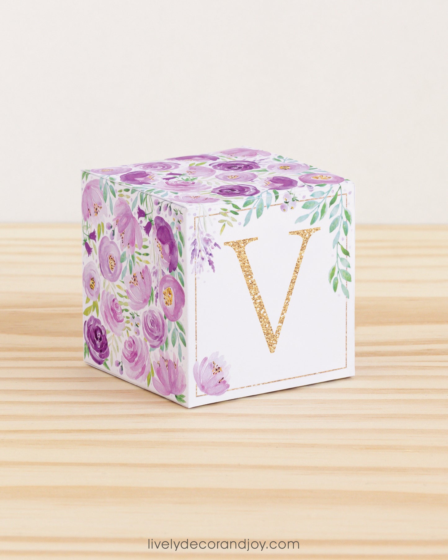Paper building block features watercolor flowers and leaves in shades of purple and green. A gold letter V is on the front.