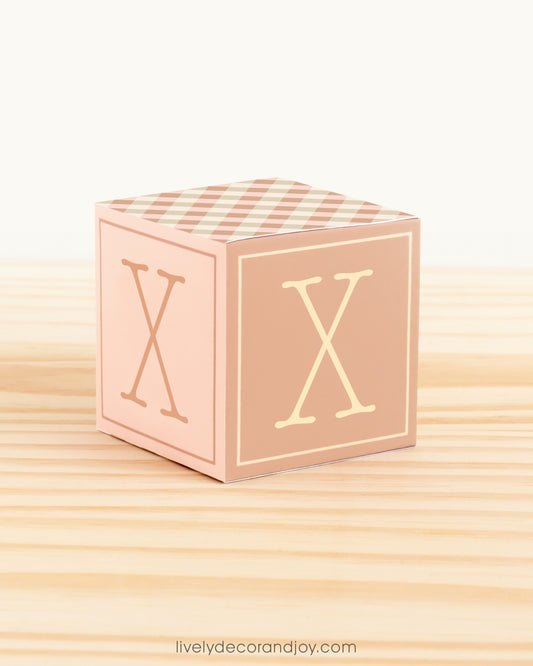 Paper building block in farmhouse colors. It has the letter X on the sides and is on a wood table or shelf.