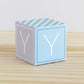 A baby block in tints of blue and light grey with the letter Y. It is on a wooden surface and is made of printed paper.