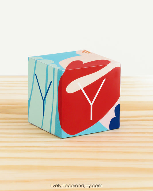 A baby block in coral reef colors with the letter Y. It is on a wooden surface and is made of printed paper.