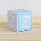 Paper building block in tints of blue and light grey. It has the letter Z on the sides and is on a wood table or shelf.