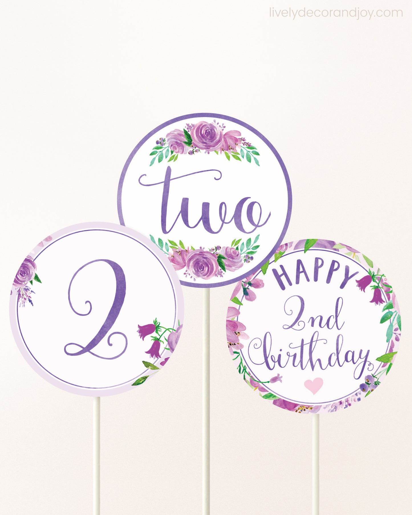 Three cupcakes toppers attached to lollipop sticks. They feature purple watercolor floral designs and purple and gold handwritten text.