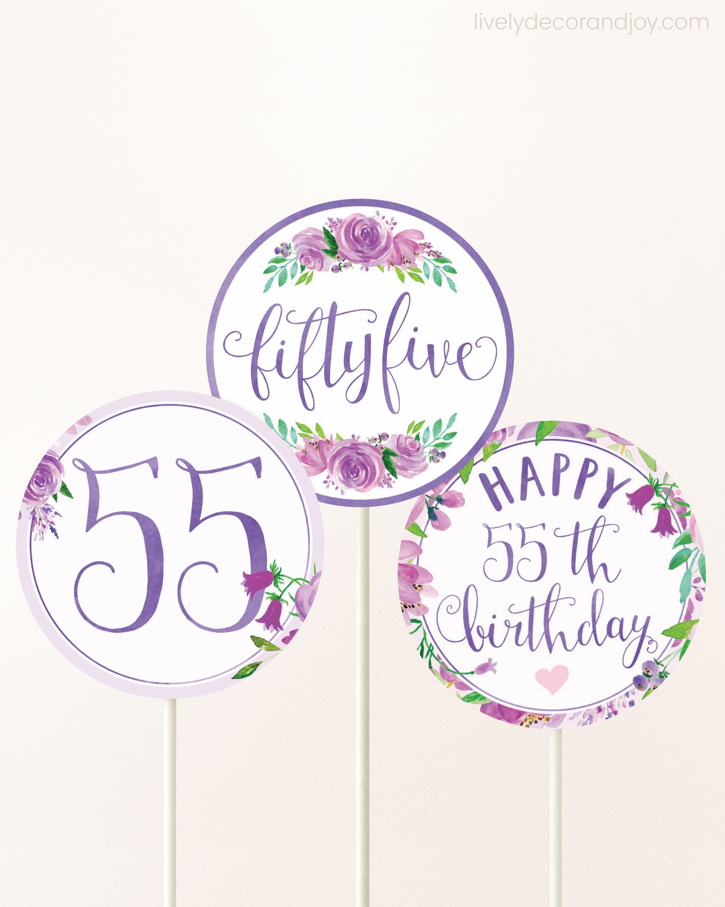 Three cupcakes toppers attached to lollipop sticks. They feature purple watercolor floral designs and purple and gold handwritten text.