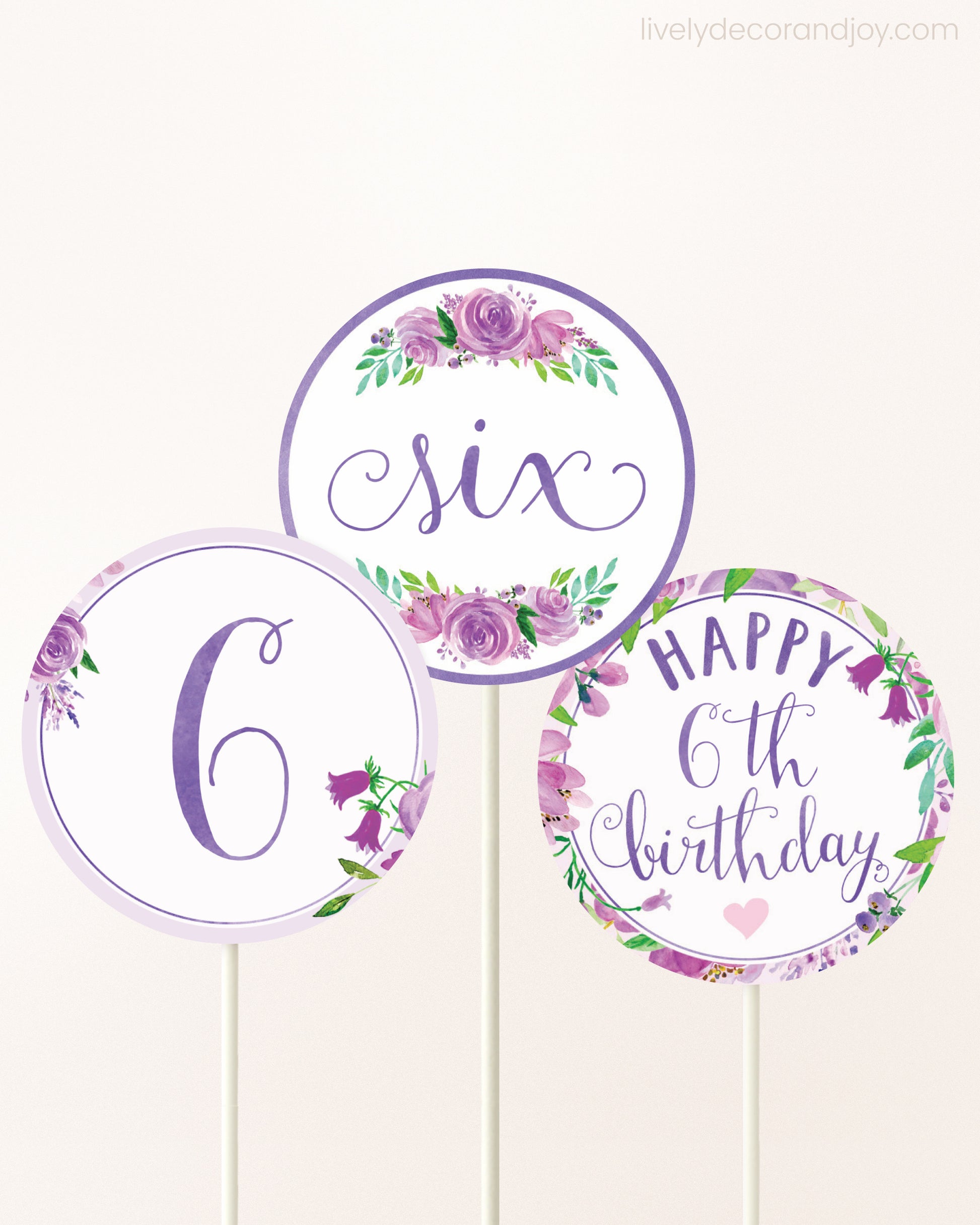 Three cupcakes toppers attached to lollipop sticks. They feature purple watercolor floral designs and purple and gold handwritten text.