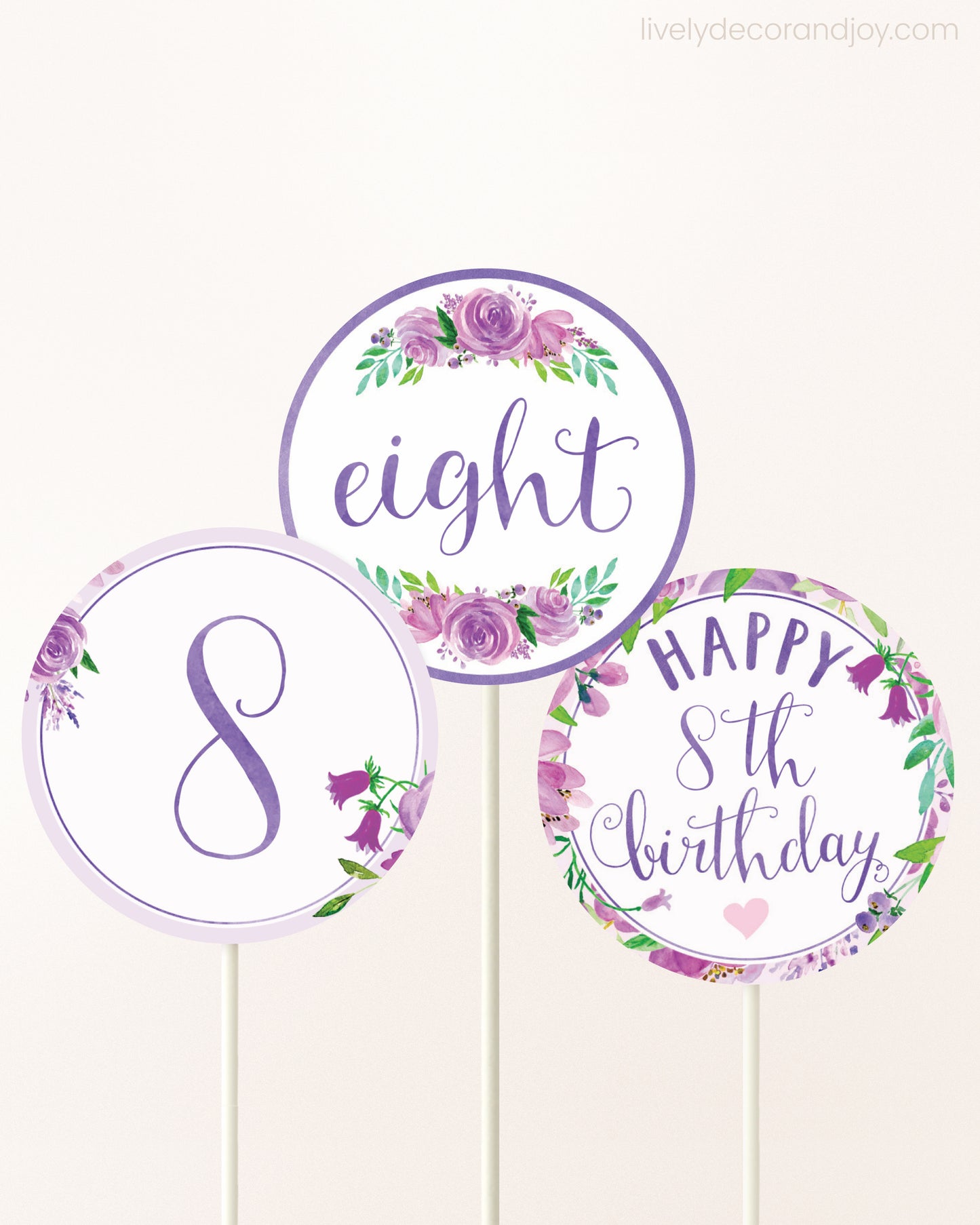 Three cupcakes toppers attached to lollipop sticks. They feature purple watercolor floral designs and purple and gold handwritten text.