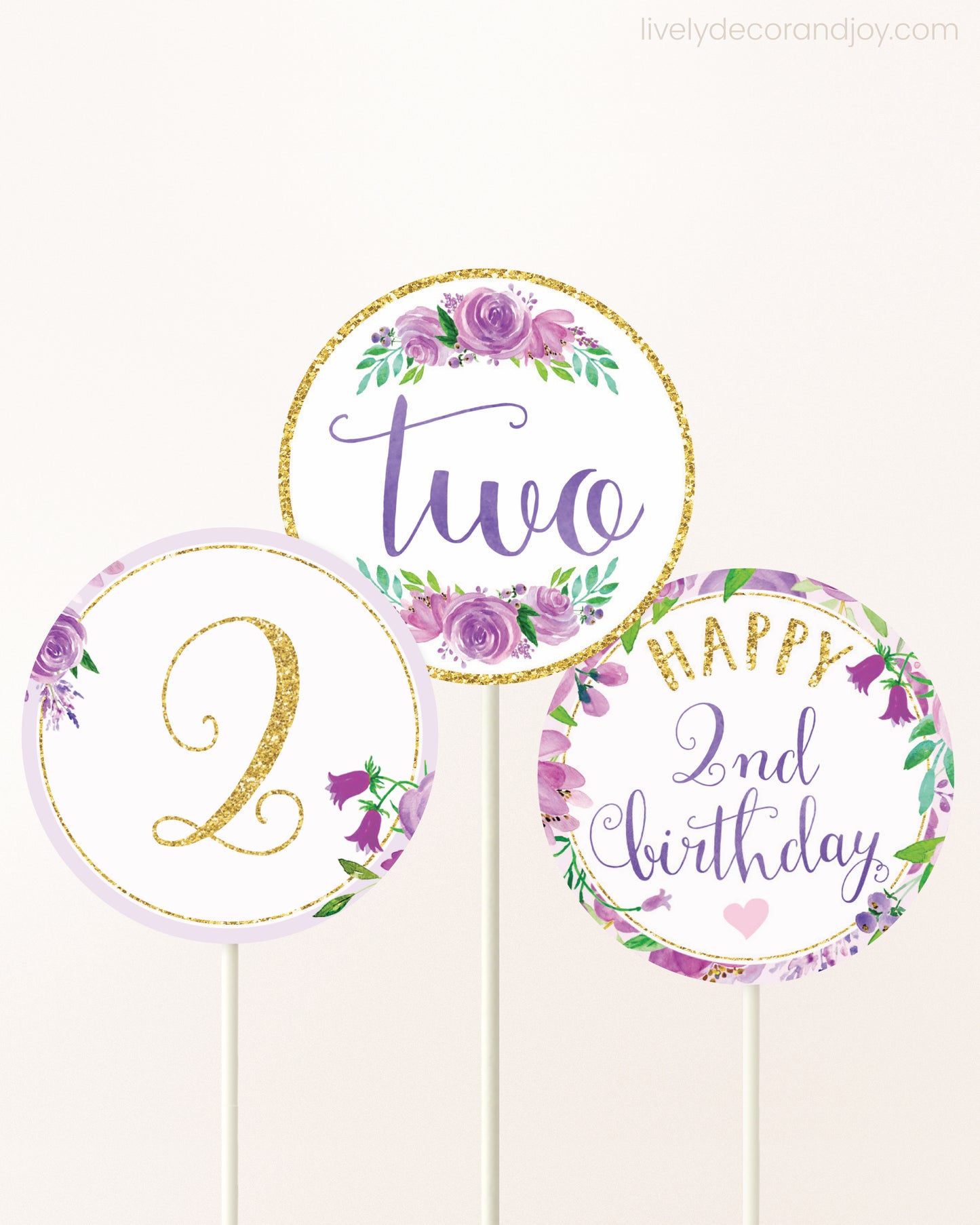 Three cupcakes toppers attached to lollipop sticks. They feature purple watercolor floral designs and purple and gold handwritten text.