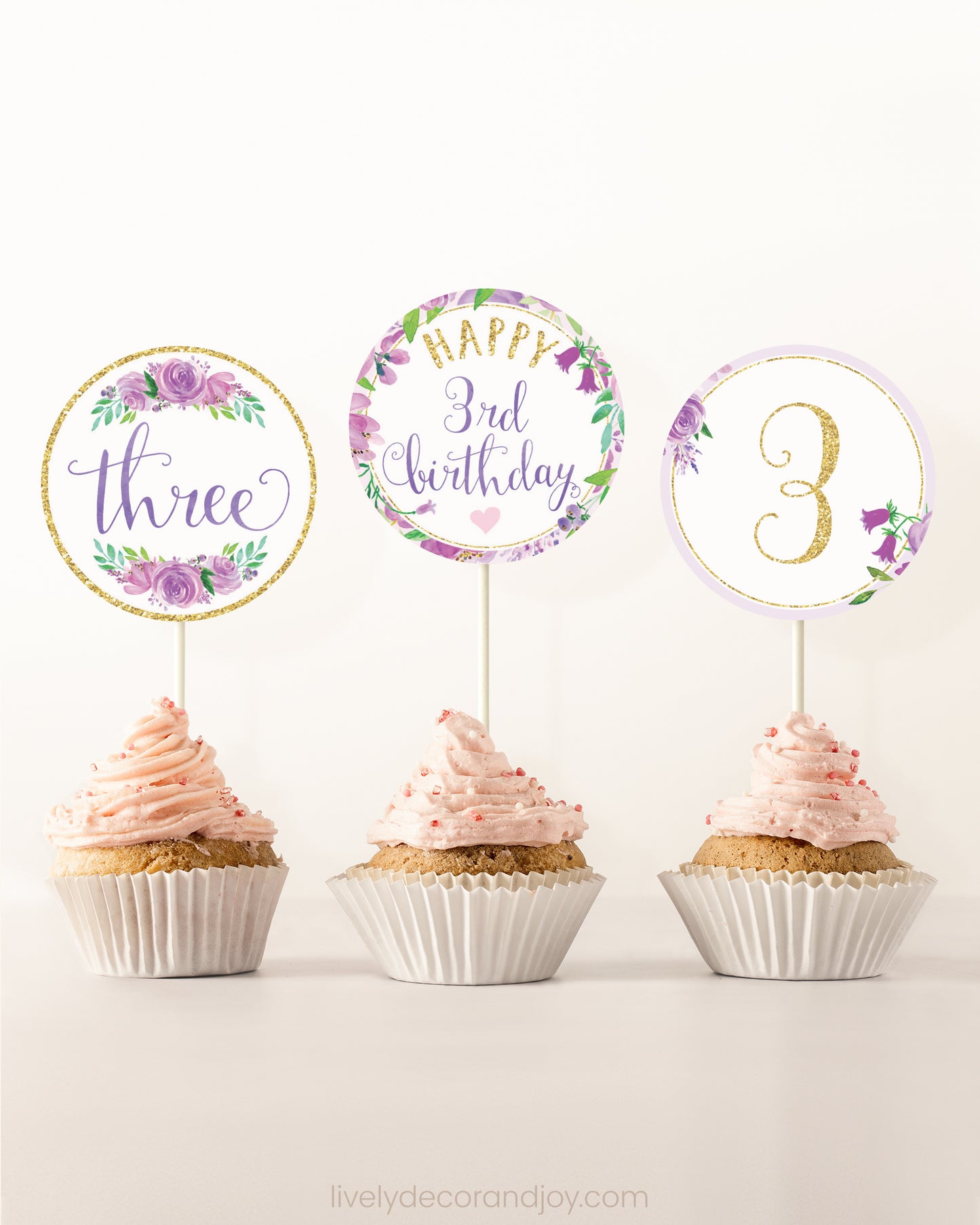 Three cupcakes side by side. Each has a round cupcake topper with purple watercolor flowers and purple and gold letters.