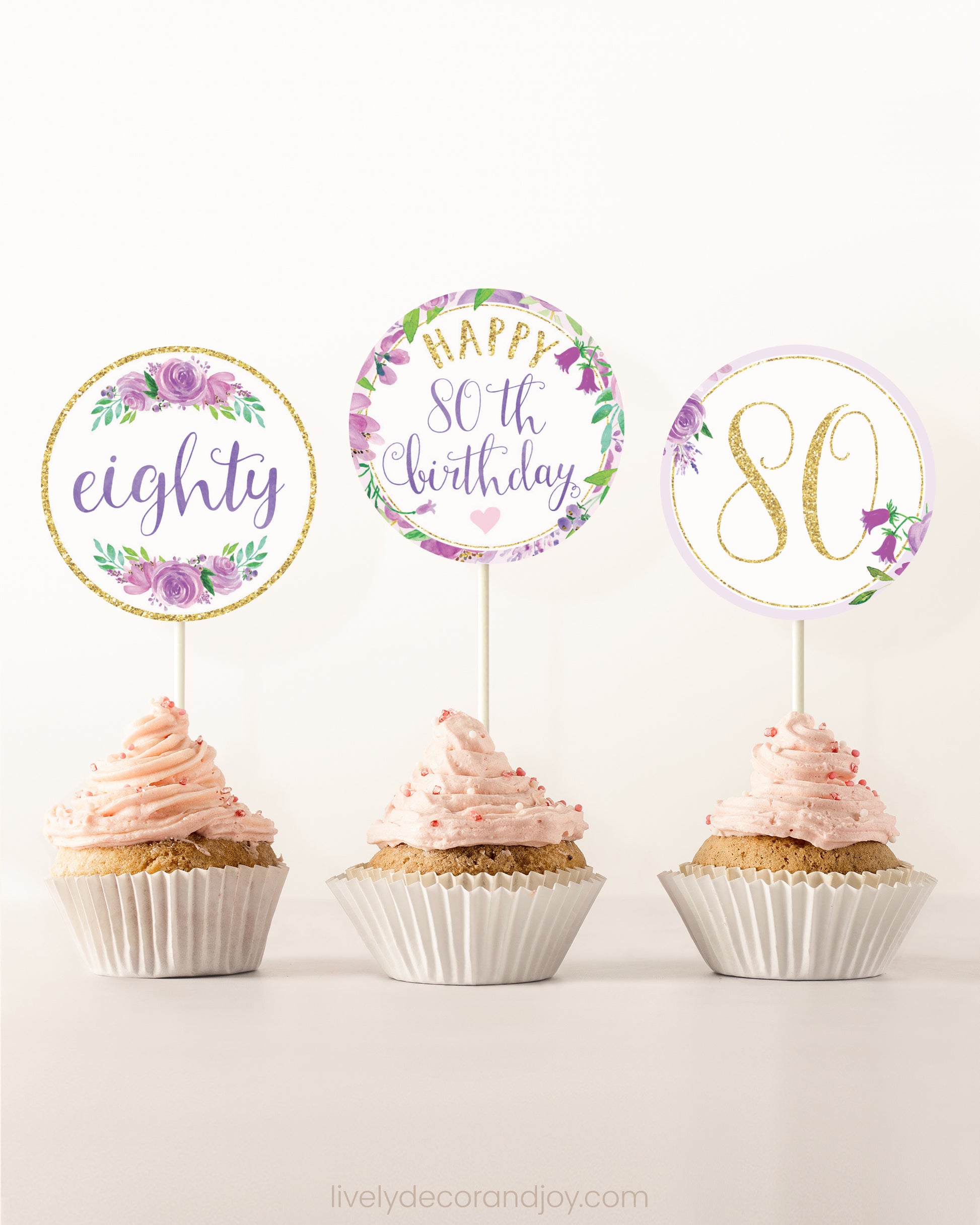 Three cupcakes side by side. Each has a round cupcake topper with purple watercolor flowers and purple and gold letters.
